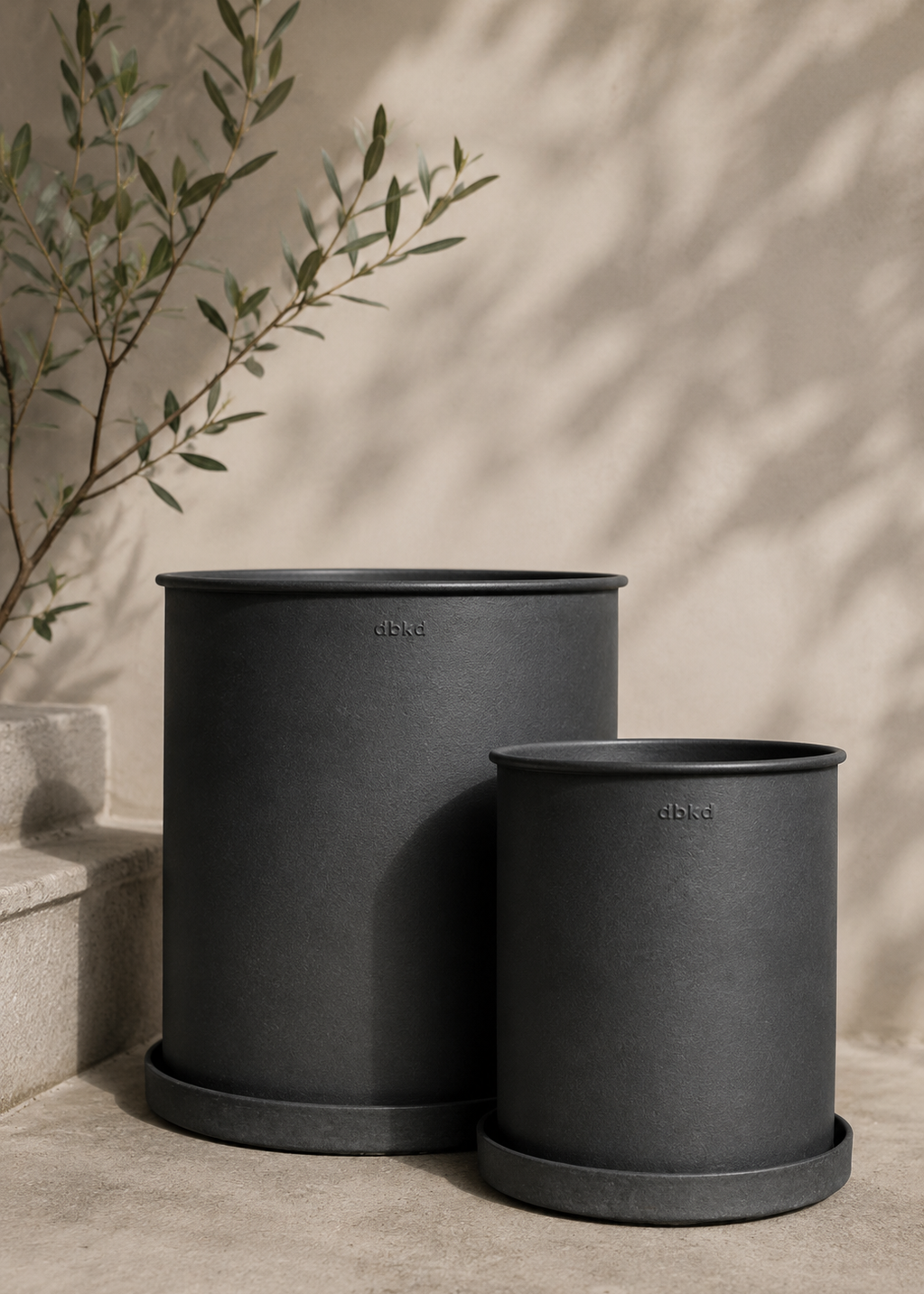 Two matte black cylindrical planters with matching saucers, one large and one small, displayed on a concrete surface with a warm beige wall and soft leaf shadows in the background.